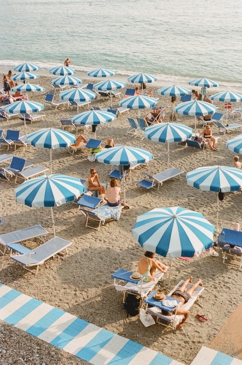 A beach is filled with people relaxing under blue and white striped umbrellas and on lounge chairs.