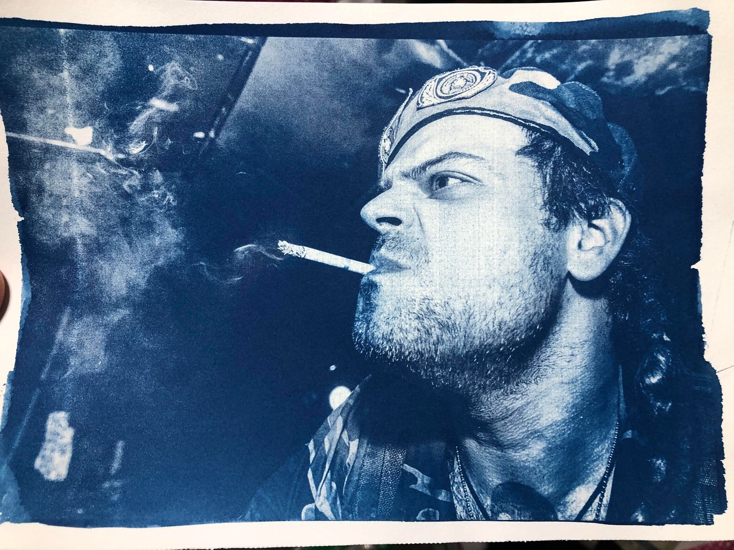 A man with a beard and a bandana smokes a cigarette, with smoke rising into the air.