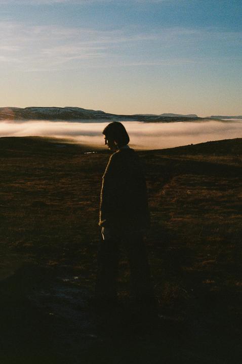A woman stands in a field with a layer of fog in the distance and mountains under a clear sky.