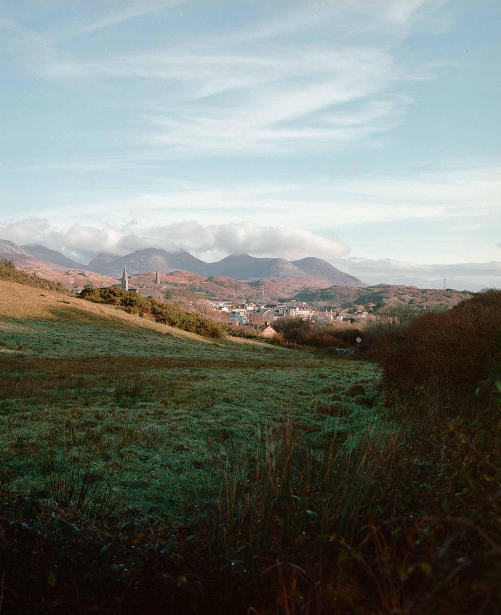 A village with two prominent spires is nestled in a valley, surrounded by rolling hills and mountains under a partly cloudy sky.