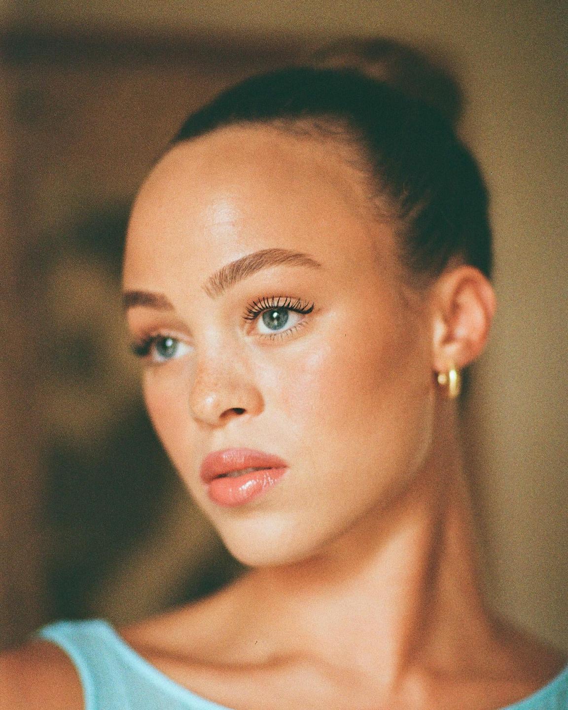 A woman with her hair in a bun and wearing a light blue top looks to the side.