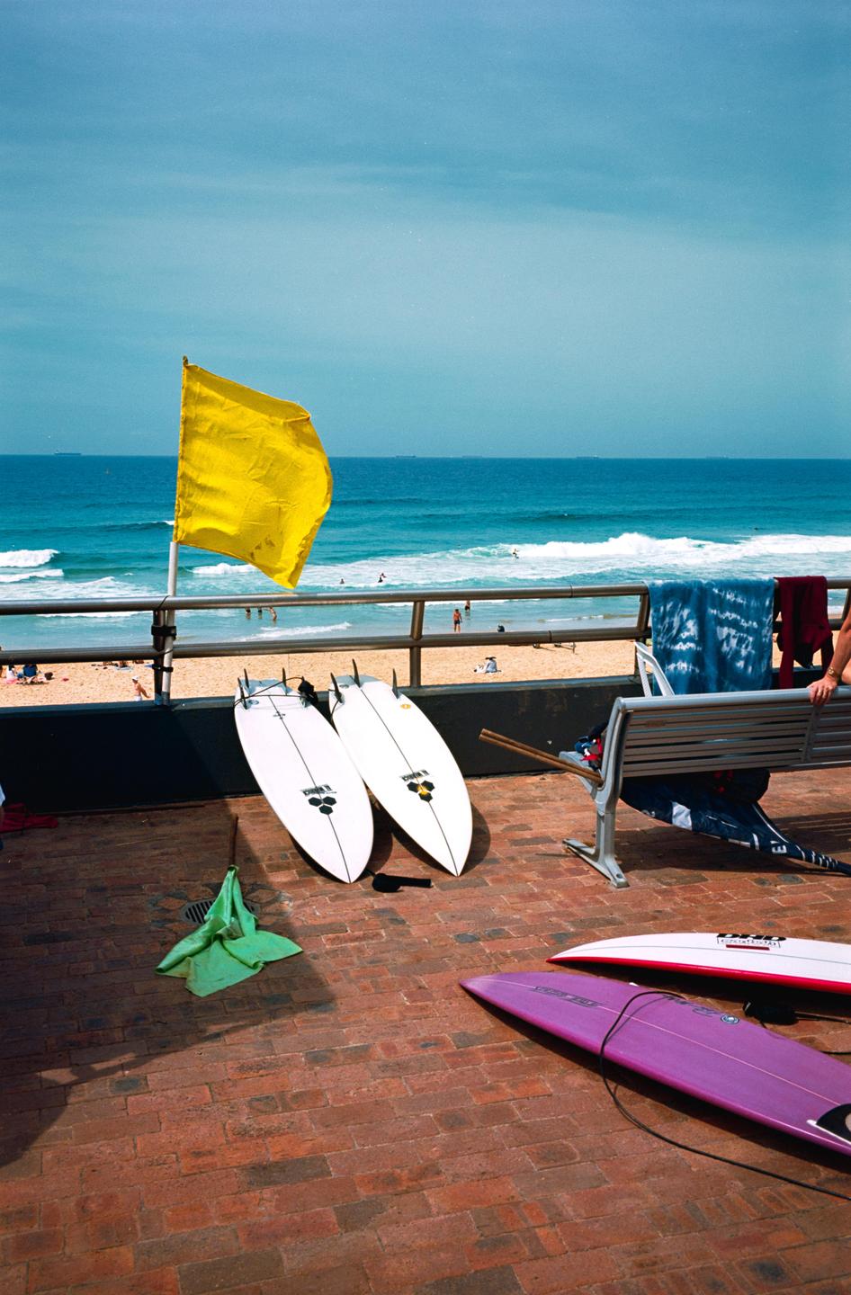 A yellow flag stands on a railing overlooking a beach with surfboards on a brick patio in the foreground.