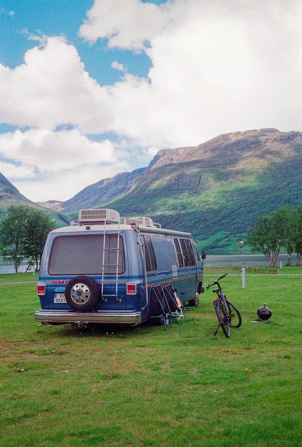 A blue GMC camper van is parked on a grassy field next to a lake with mountains in the background.