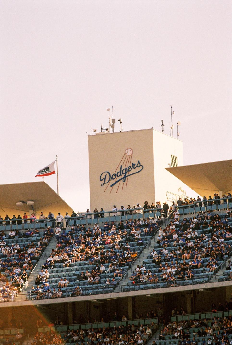 A large crowd of people sits in the stands of a baseball stadium with the Dodgers logo visible on a building.