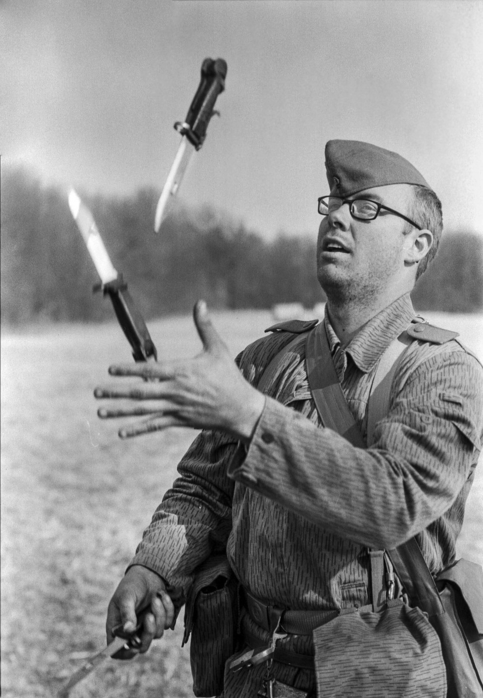 A man in military uniform and glasses juggles three bayonets in an open field.