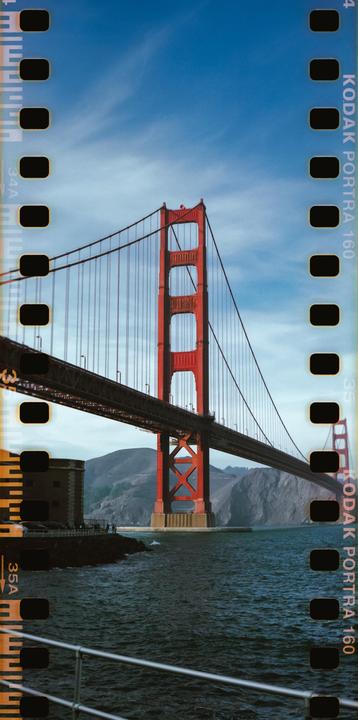 The Golden Gate Bridge spans across a body of water with mountains in the background under a blue sky.