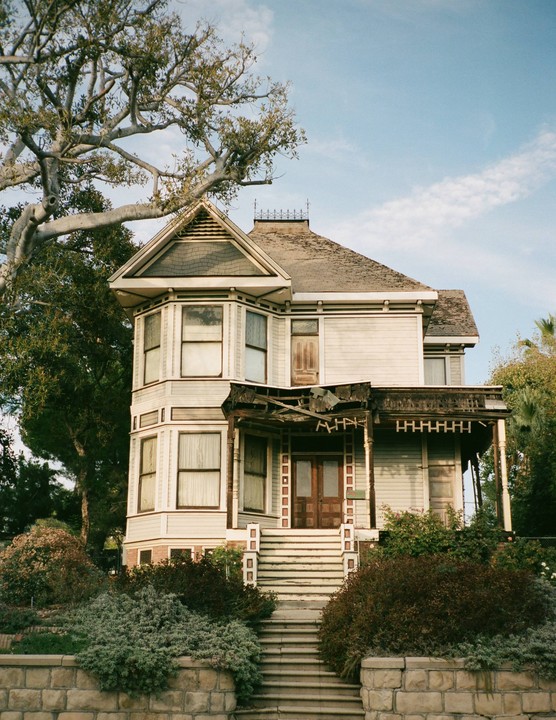 A large, light-colored house with a damaged porch and bay windows stands behind a stone wall and overgrown bushes.