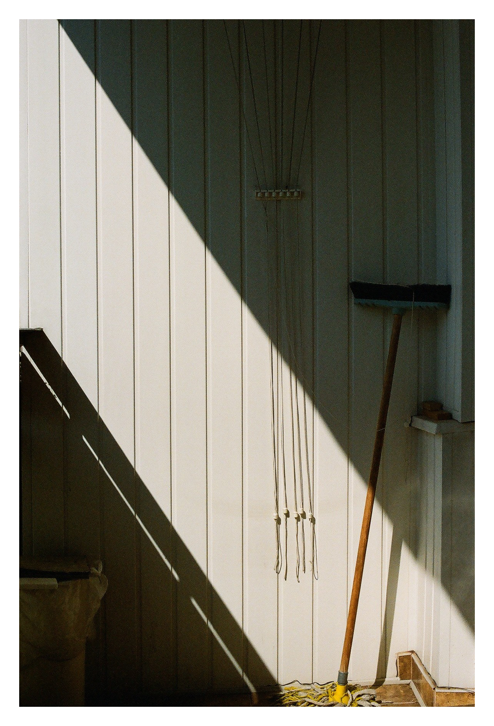 A broom and a clothesline hang on a white wall, with a strong diagonal shadow cutting across the frame.