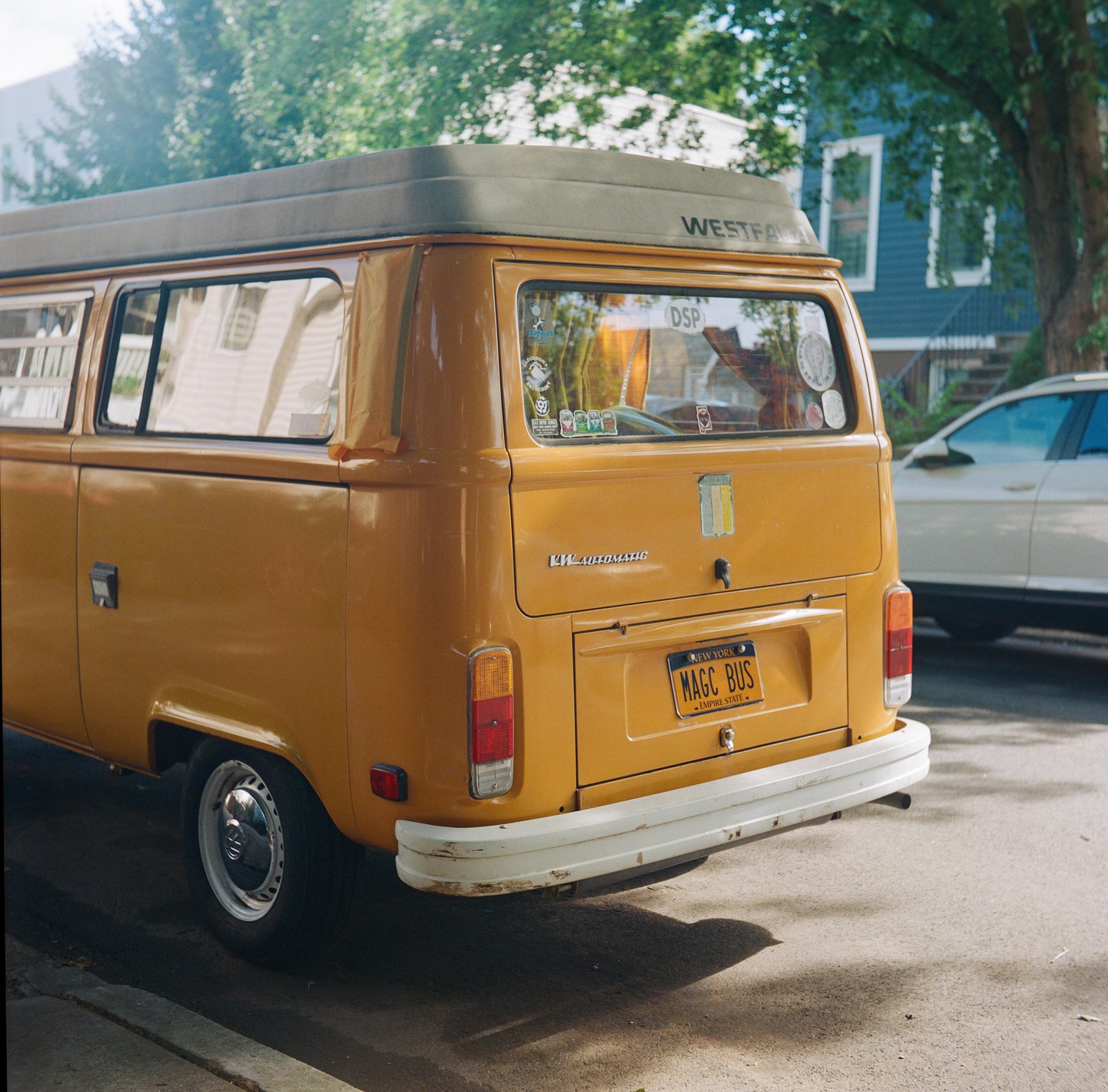A yellow van with a white bumper is parked on a street next to a tree and a house.