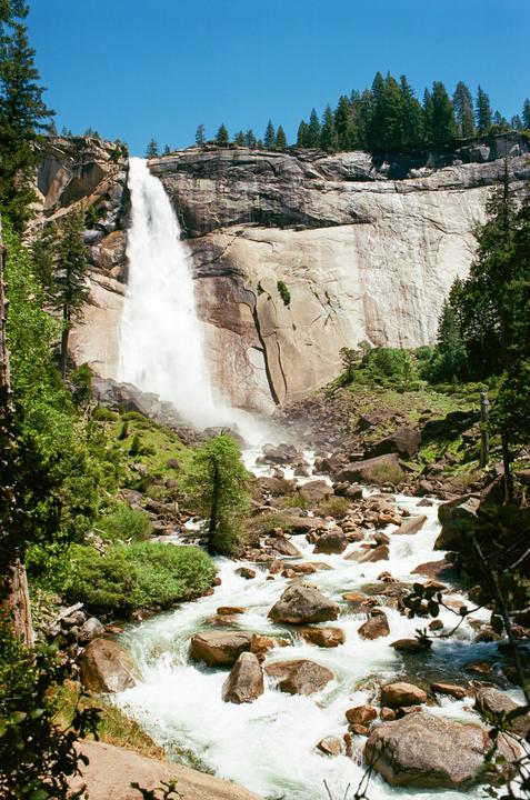 A large waterfall cascades down a rocky cliff into a river filled with boulders and surrounded by green trees.