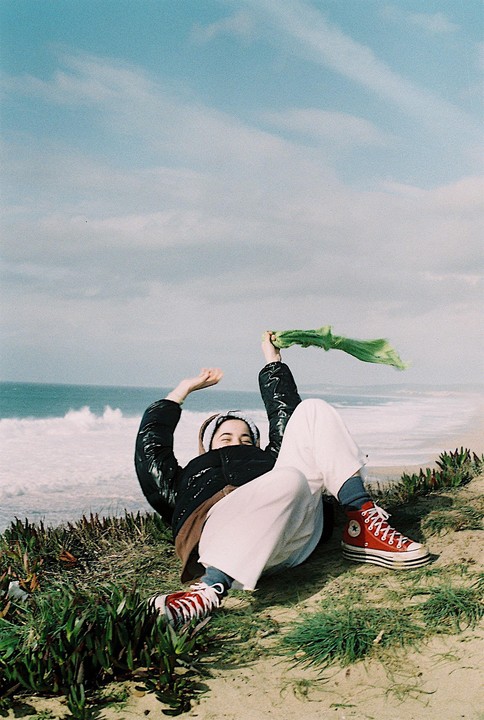 A woman in a black jacket and white pants is lying on a sandy cliff overlooking the ocean, holding a green scarf.