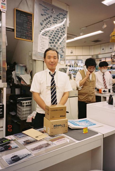 A man in a white shirt and striped tie stands behind a counter with two boxes labeled "Nikon S3" in a store.