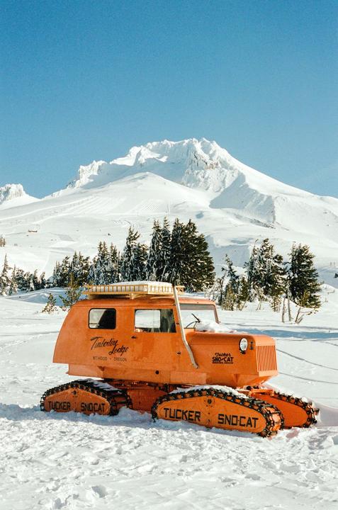 An orange snowcat is parked in the snow with a mountain and trees in the background.