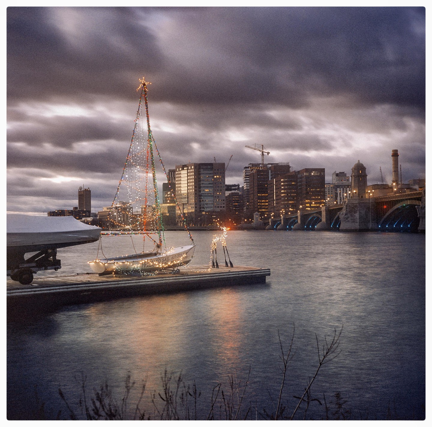 A small sailboat decorated with Christmas lights is docked in a river with a city skyline and bridge in the background under a cloudy sky.