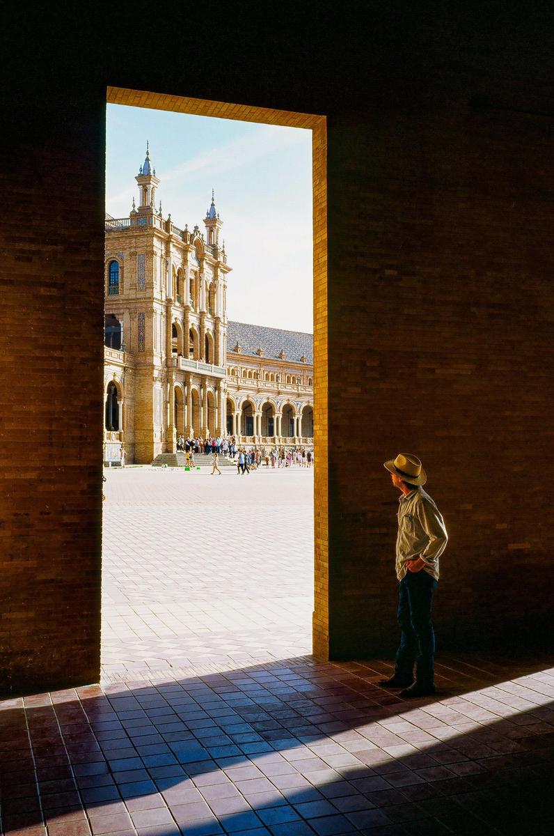 A man in a hat stands in shadow looking out through a doorway at a large ornate building and people in a sunny plaza.