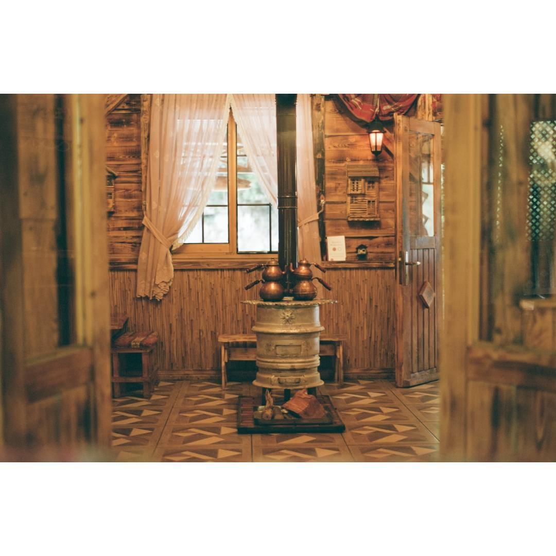 A rustic room with wooden walls and floor features a central stove with teapots on top, a window with sheer curtains, and an open door.