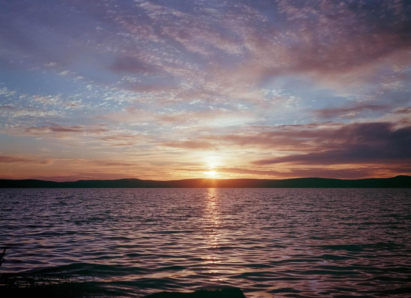 The sun sets over a body of water with a distant shoreline and colorful clouds.