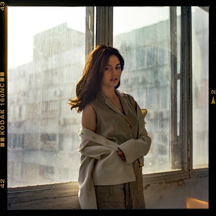A woman with long hair poses in front of a large, dirty window, wearing a sleeveless top and a jacket draped over her shoulders.