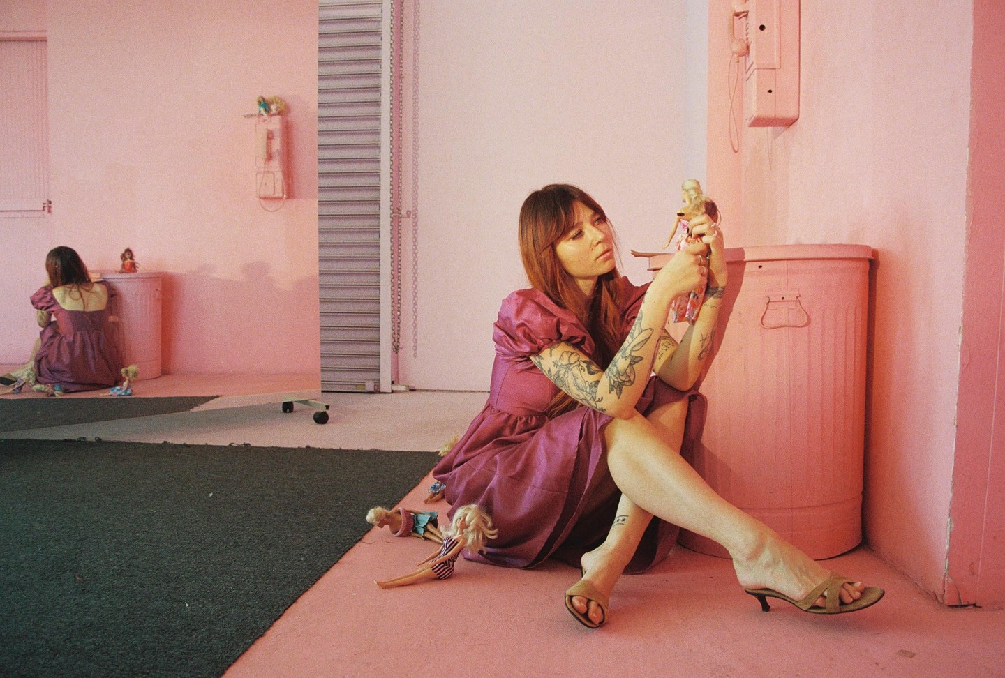 A woman in a purple dress sits on a pink floor, holding a doll, with another woman in the background and several dolls scattered around.