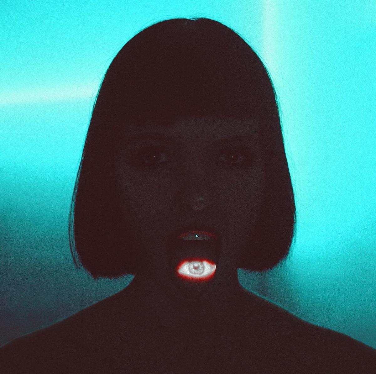 A woman with dark hair and a bob haircut has an eye projected onto her open mouth, with a bright blue background.
