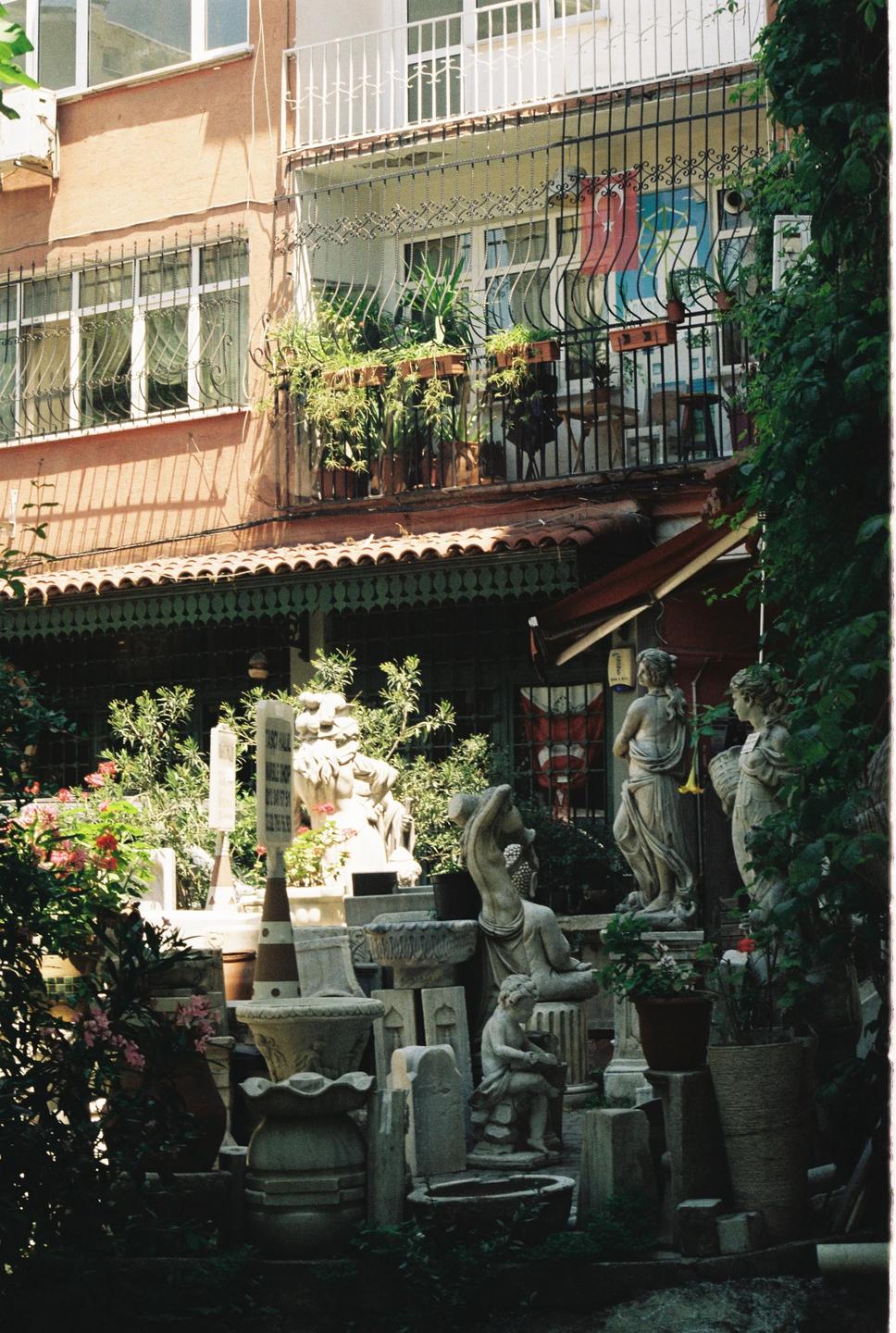 A collection of statues and planters are arranged in front of a building with balconies and lush greenery.