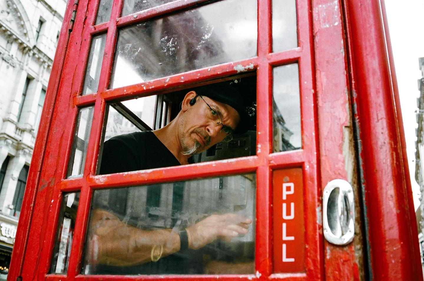 A man with a goatee and glasses looks out from a red telephone booth.