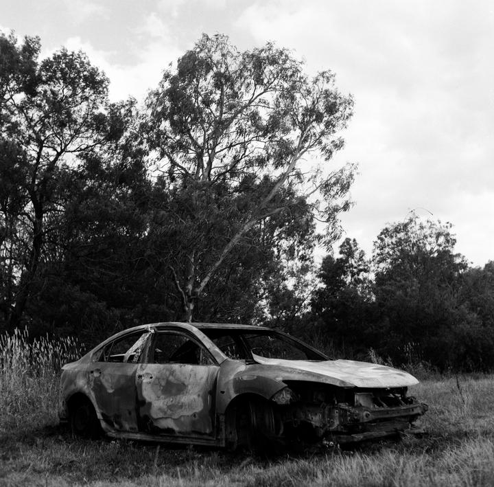 A burnt car sits in a grassy field with trees in the background under a cloudy sky.