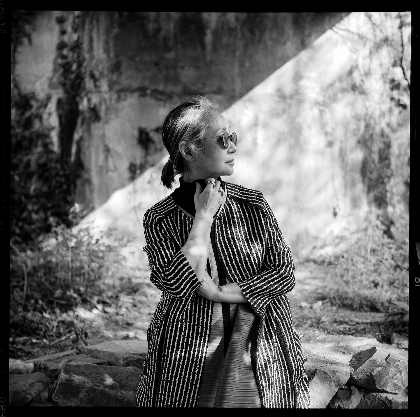 A woman wearing sunglasses and a striped coat stands outdoors with her hand to her neck.