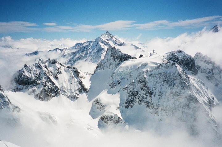 Snow-covered mountains rise above a sea of clouds under a blue sky.