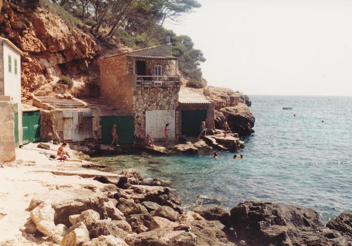 People are swimming and relaxing on a rocky beach with stone buildings built into a cliff.