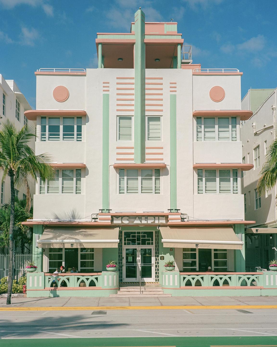 A pastel-colored Art Deco building with awnings and palm trees stands beside a street.