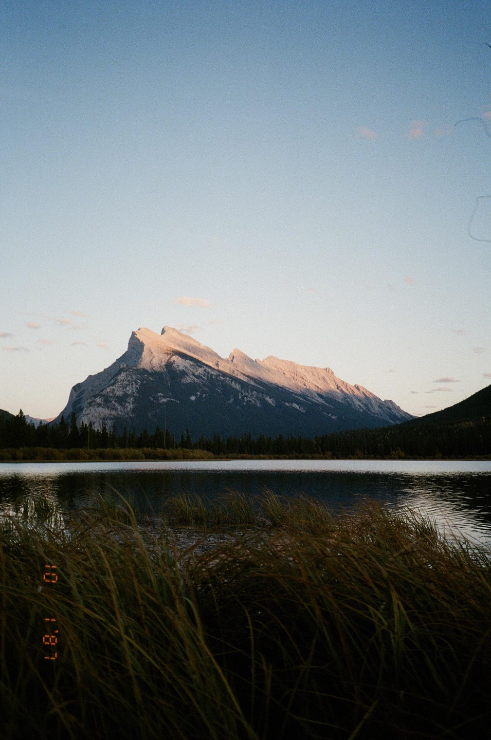 A mountain range with a prominent peak is reflected in a lake with tall grass in the foreground.