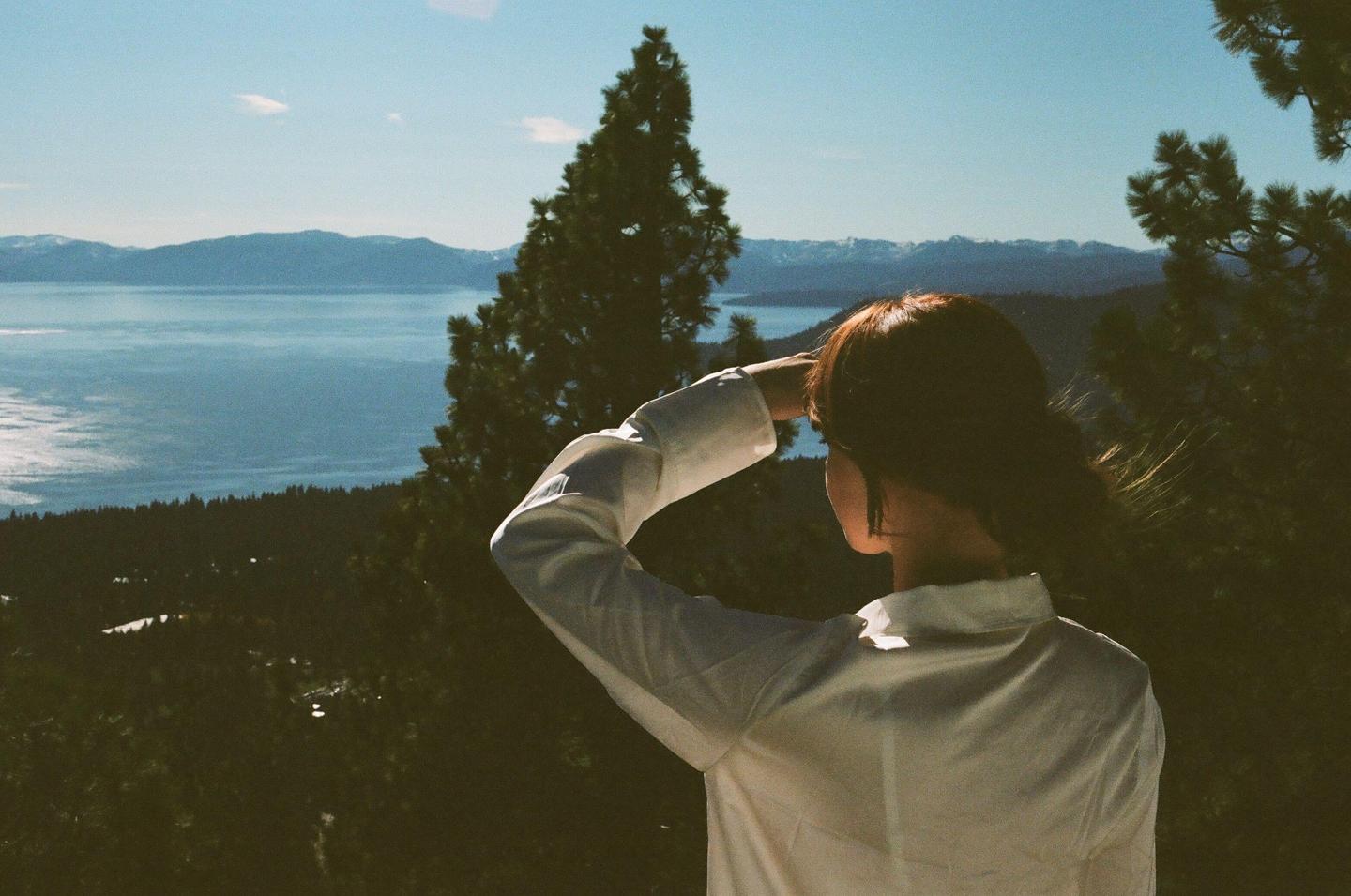 A woman in a white shirt looks out over a large lake surrounded by mountains and trees.