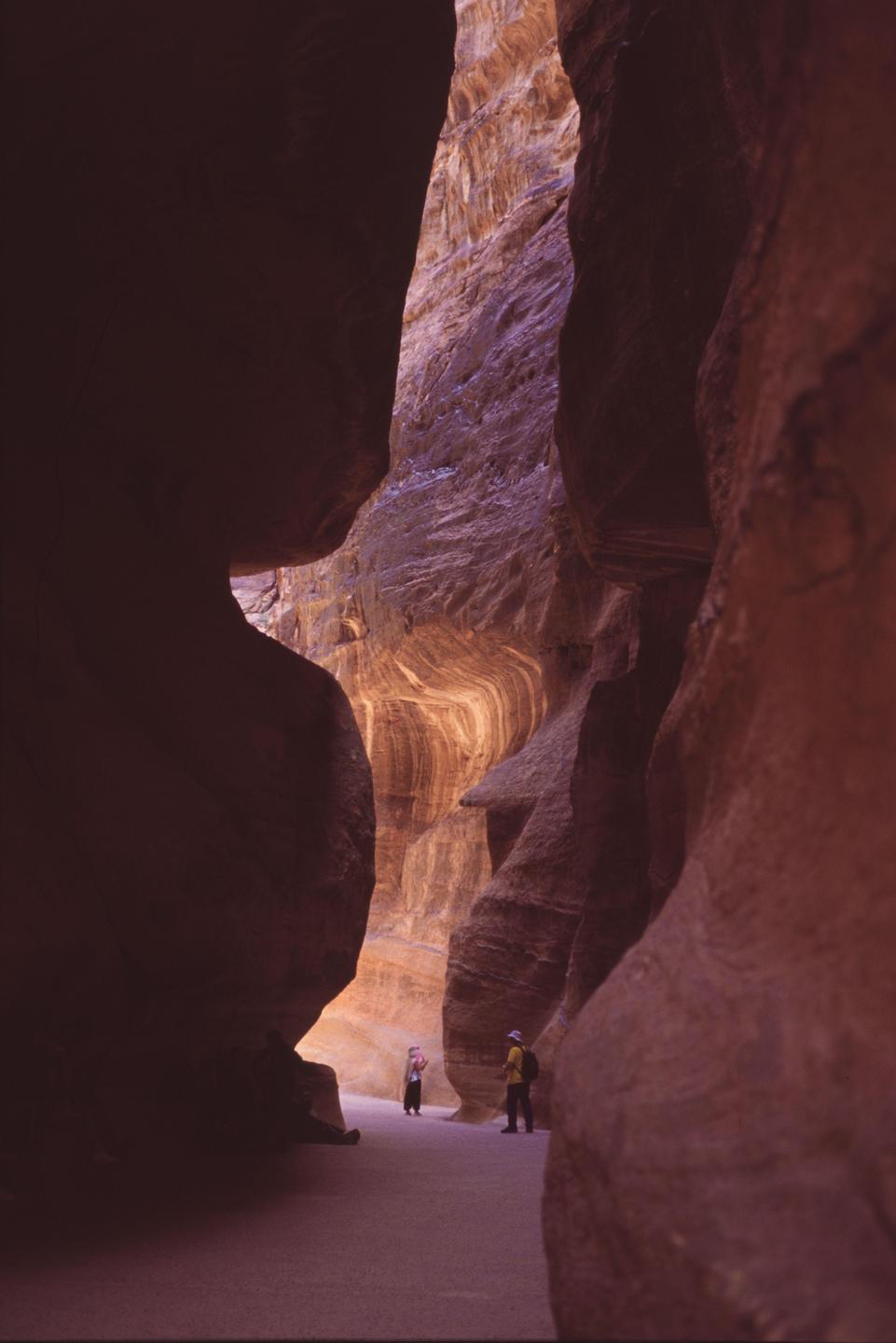 Two people stand in a narrow canyon with towering rock walls illuminated by sunlight.