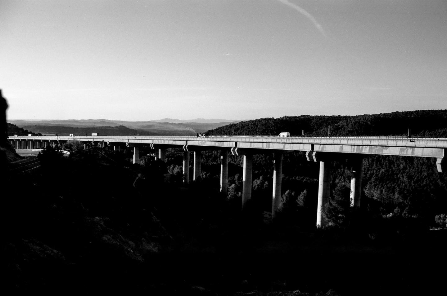 A long highway bridge with several vehicles crosses a valley with mountains in the distance.
