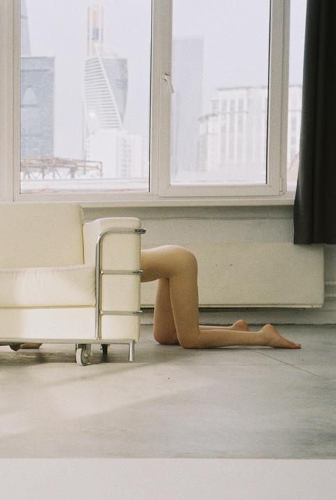 A person is kneeling on the floor behind a white sofa, with a city visible through a window in the background.