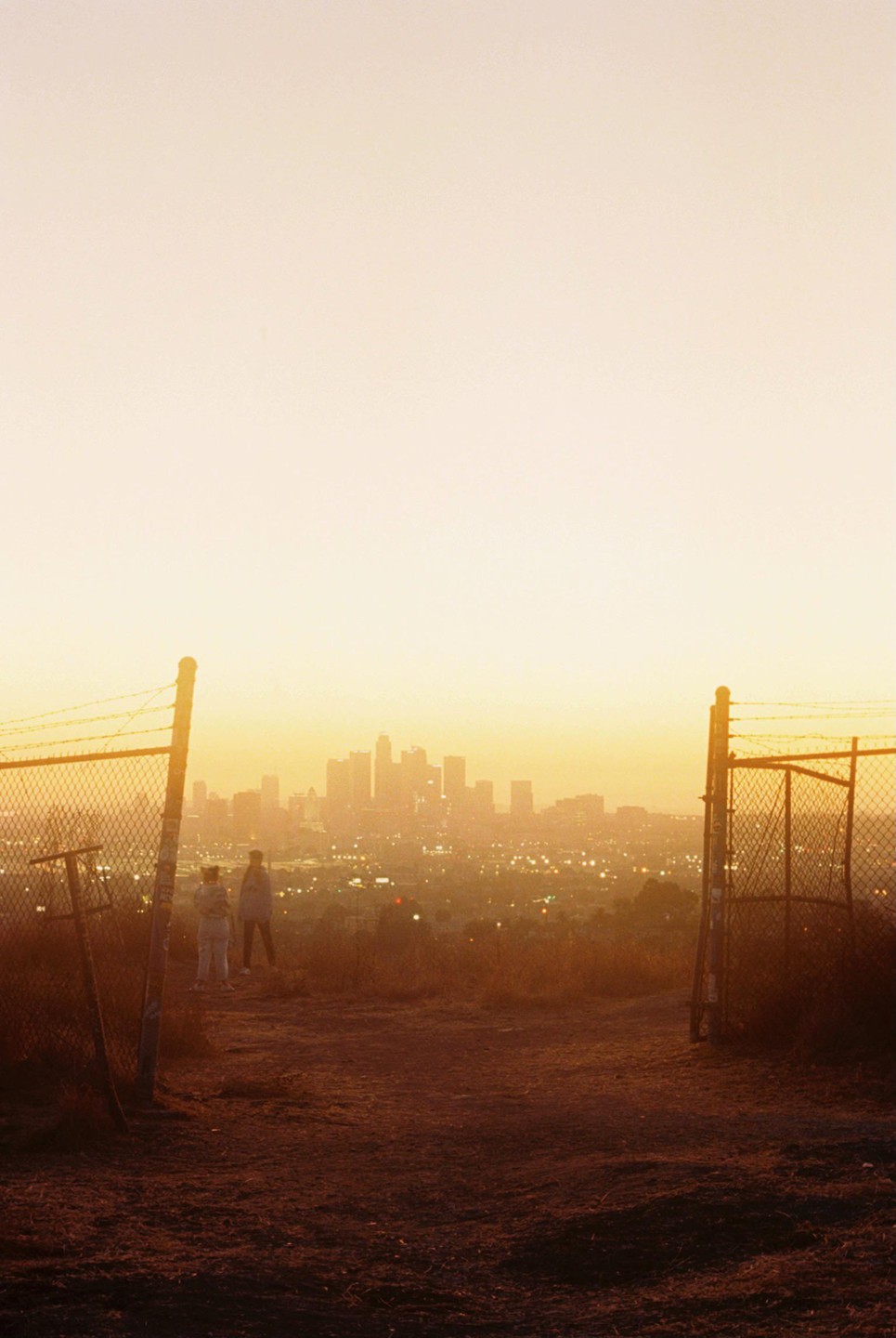 Two people stand on a hill overlooking a city skyline at sunset, framed by two chain-link fence posts.