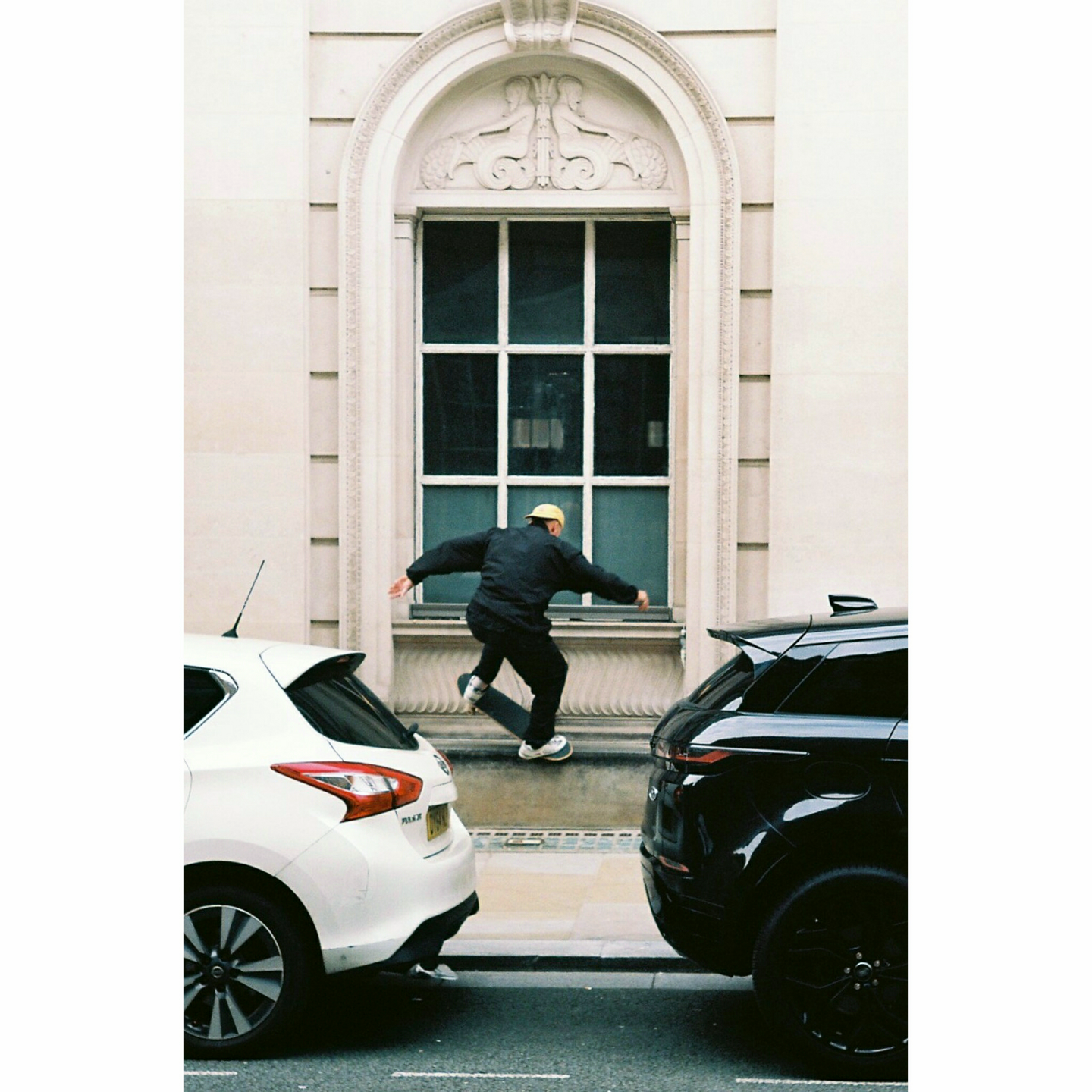 A skateboarder performs a trick in front of a large arched window between two parked cars.