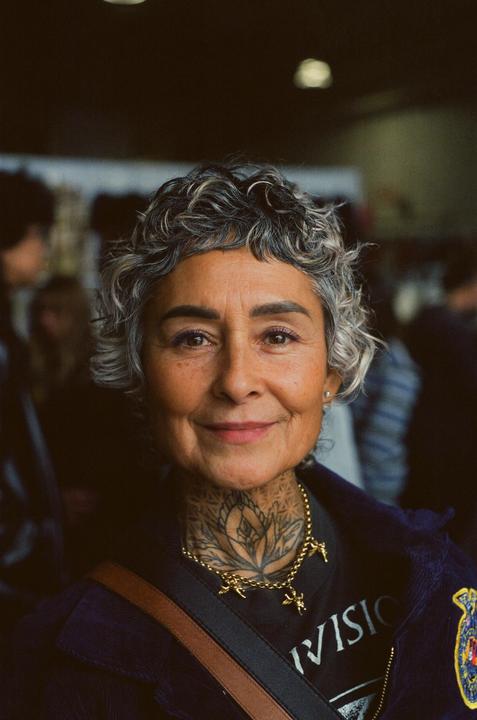 A woman with short curly hair and a tattooed neck smiles at the viewer.