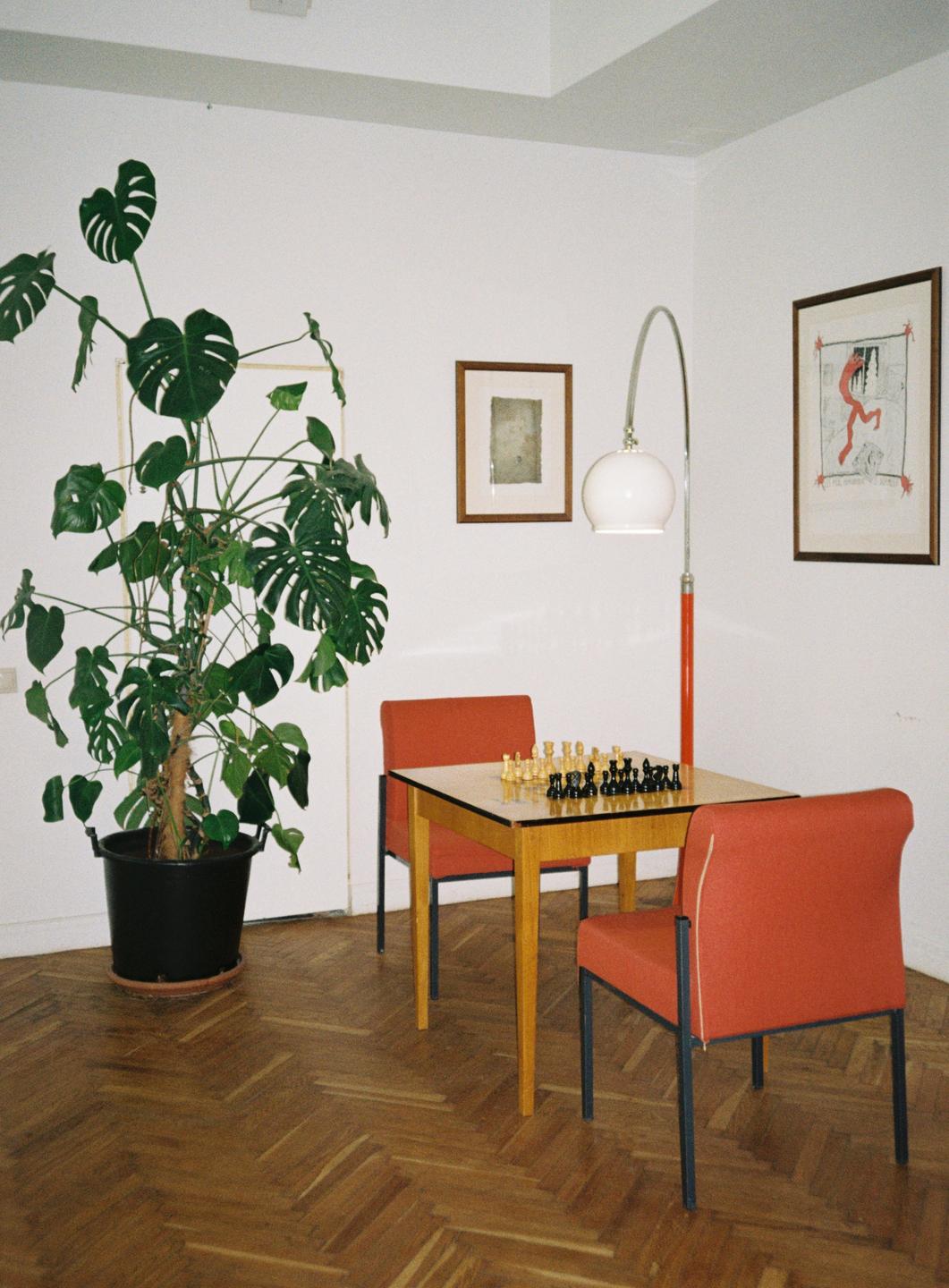 A monstera plant in a black pot stands next to a small wooden table with a chessboard and two orange chairs.
