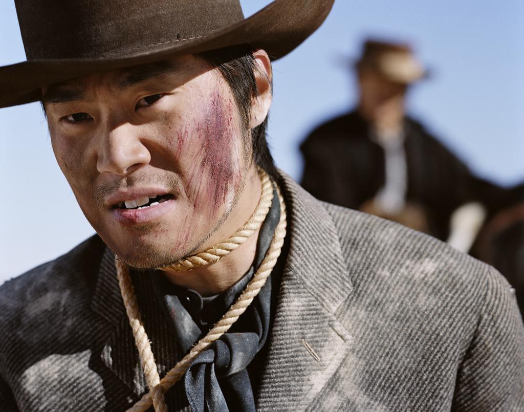 A man with a rope around his neck and a bloody face wears a cowboy hat and a jacket.