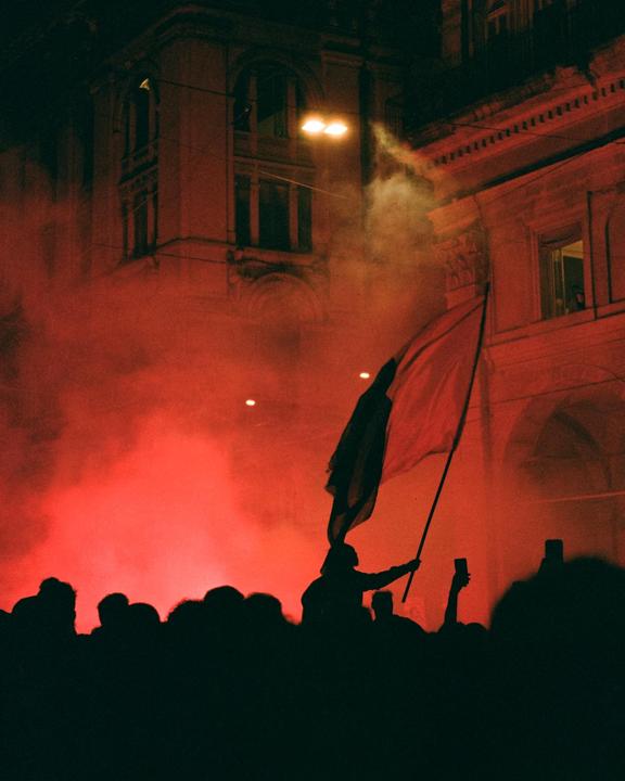 A crowd of people are silhouetted against a red smoky background with a building in the distance.
