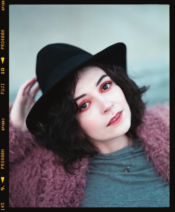 A woman wearing a black hat and a pink fluffy coat looks directly at the viewer.