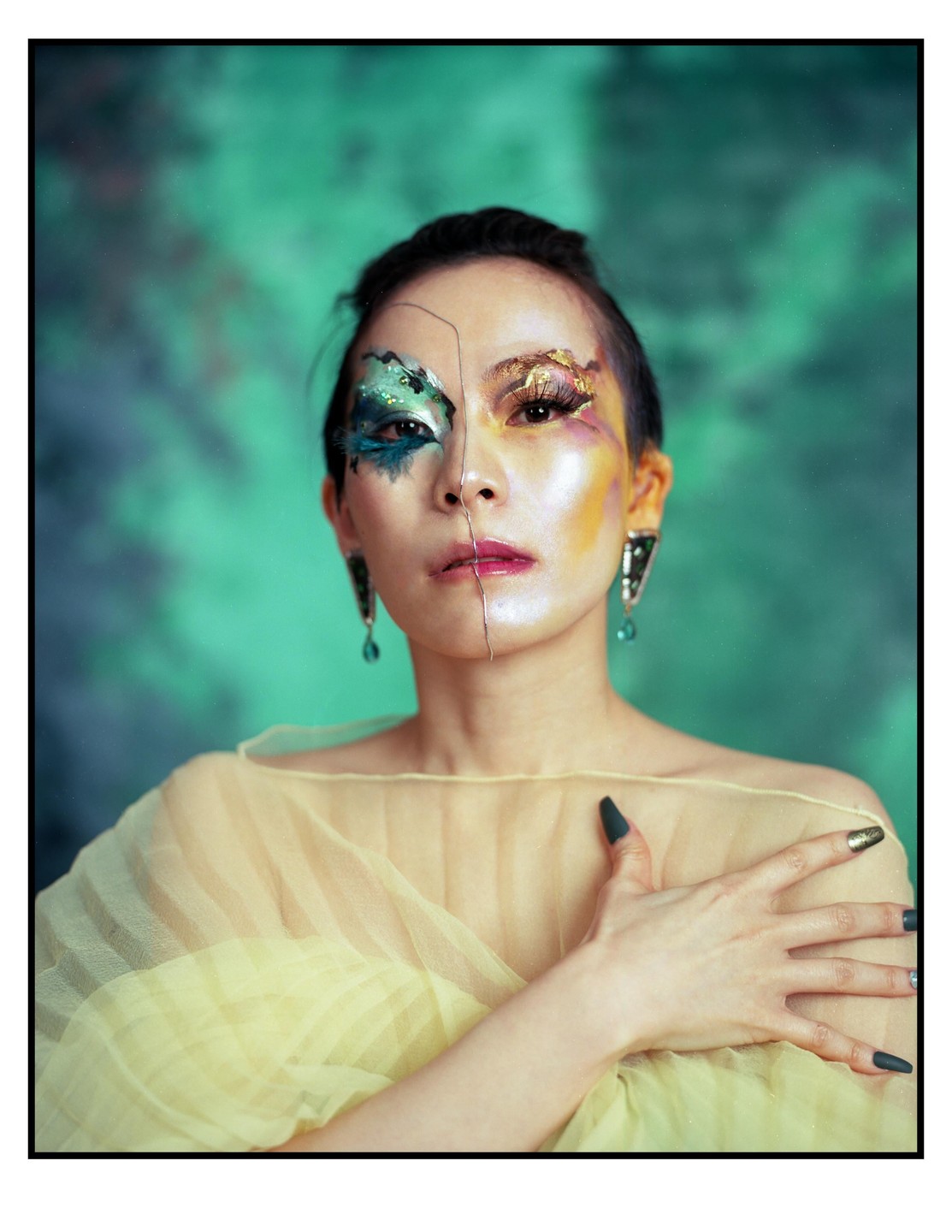 A woman with elaborate, colorful makeup on her face and a sheer yellow garment poses against a green background.