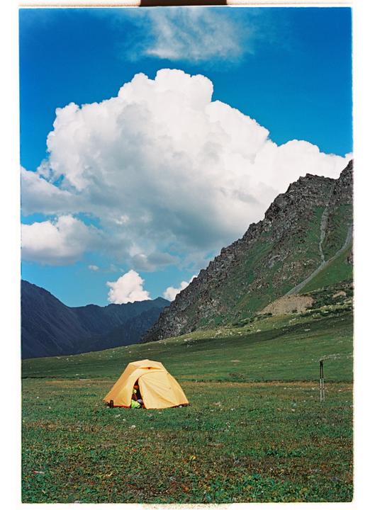 A yellow tent sits in a green field with mountains and a blue sky with clouds in the background.