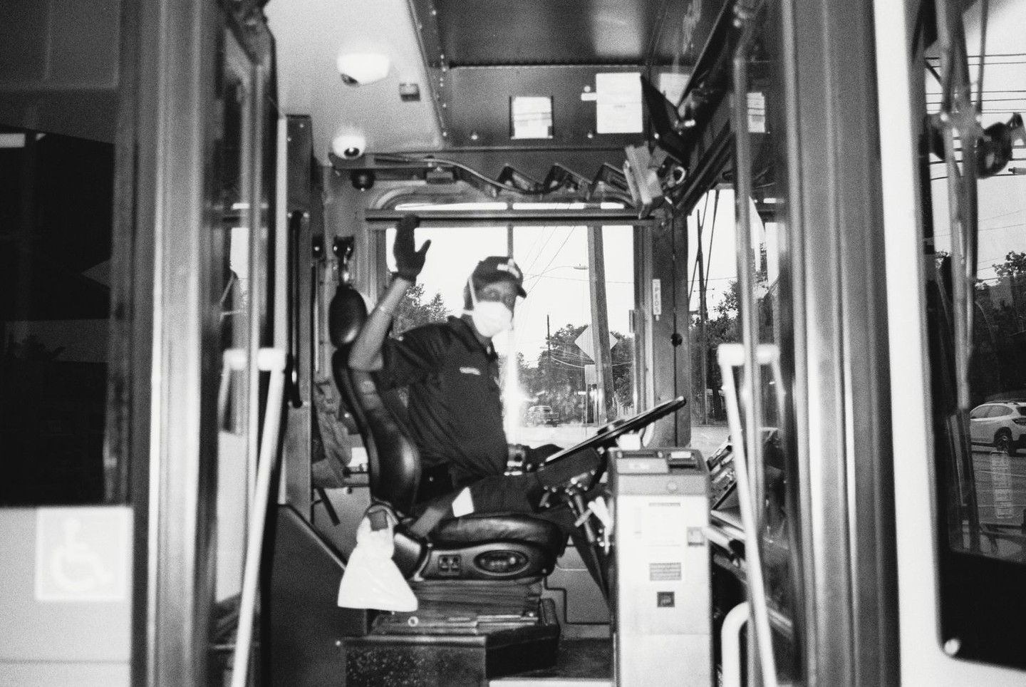 A bus driver wearing a face mask and cap waves from the driver's seat of a bus.