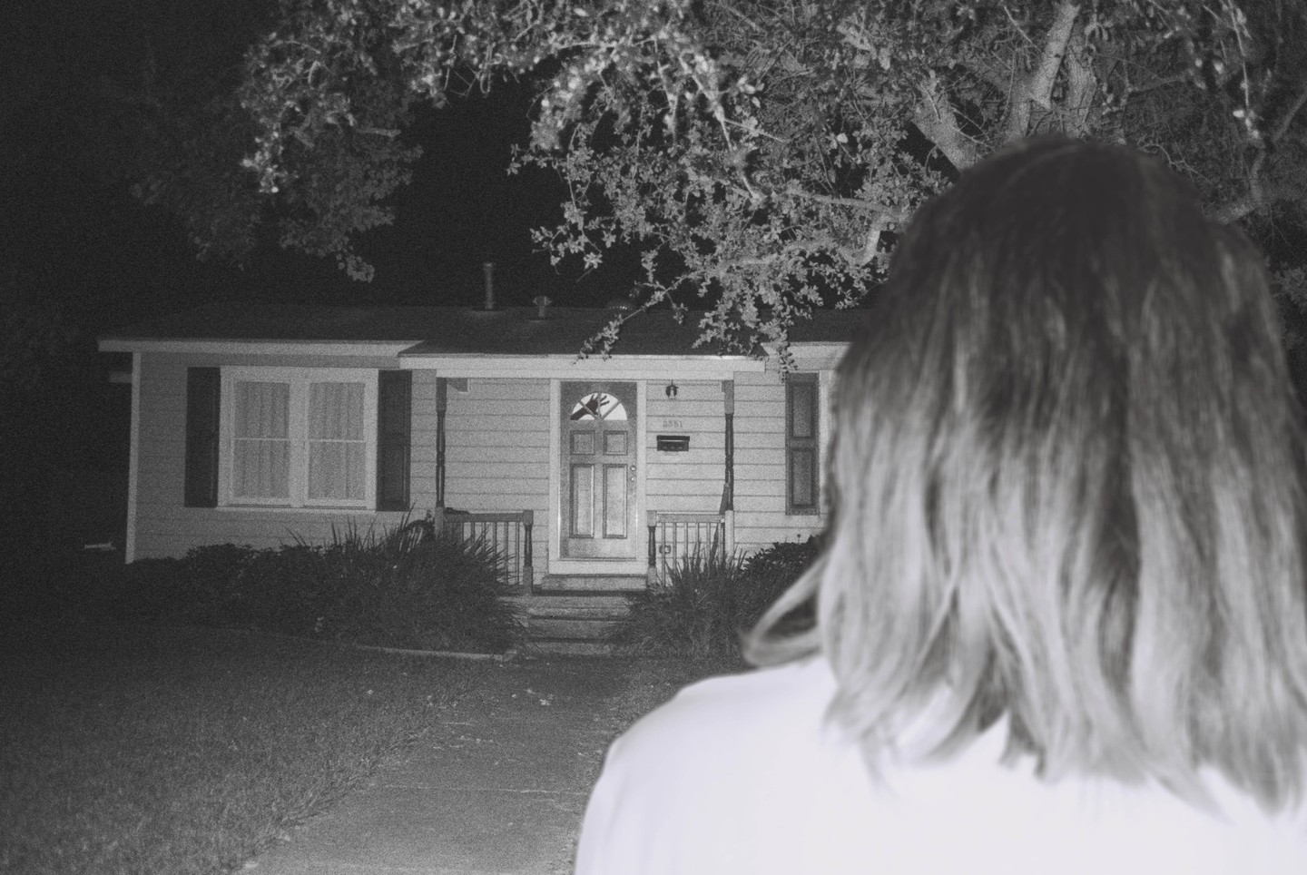 A person with long hair stands with their back to the camera, looking at a house at night.