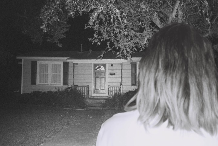 A person with long hair stands with their back to the camera, looking at a house at night.