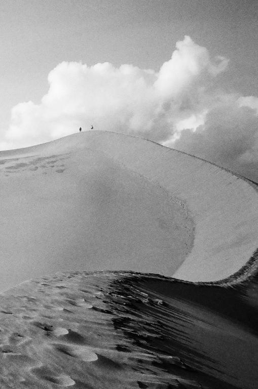 Two people stand on the crest of a large sand dune under a cloudy sky.