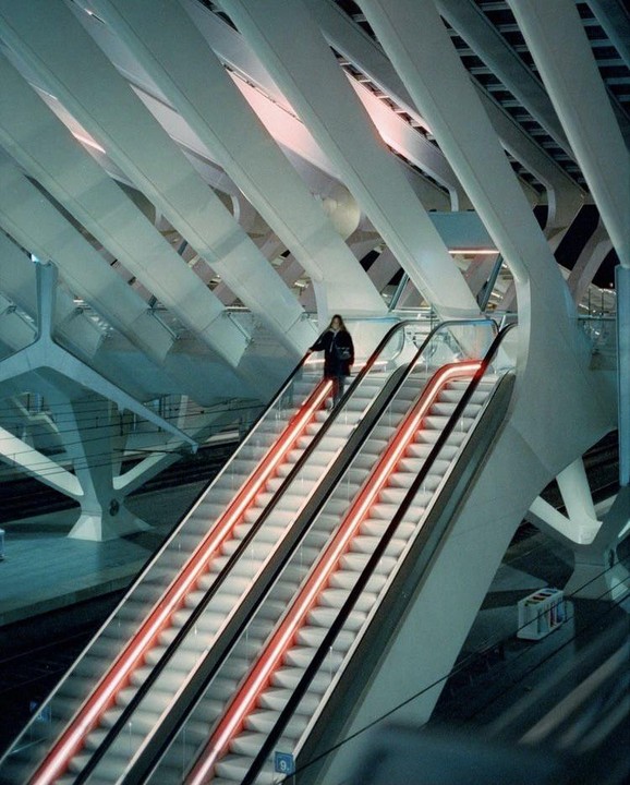 A woman rides an escalator with red neon lights in a modern building with a high ceiling.